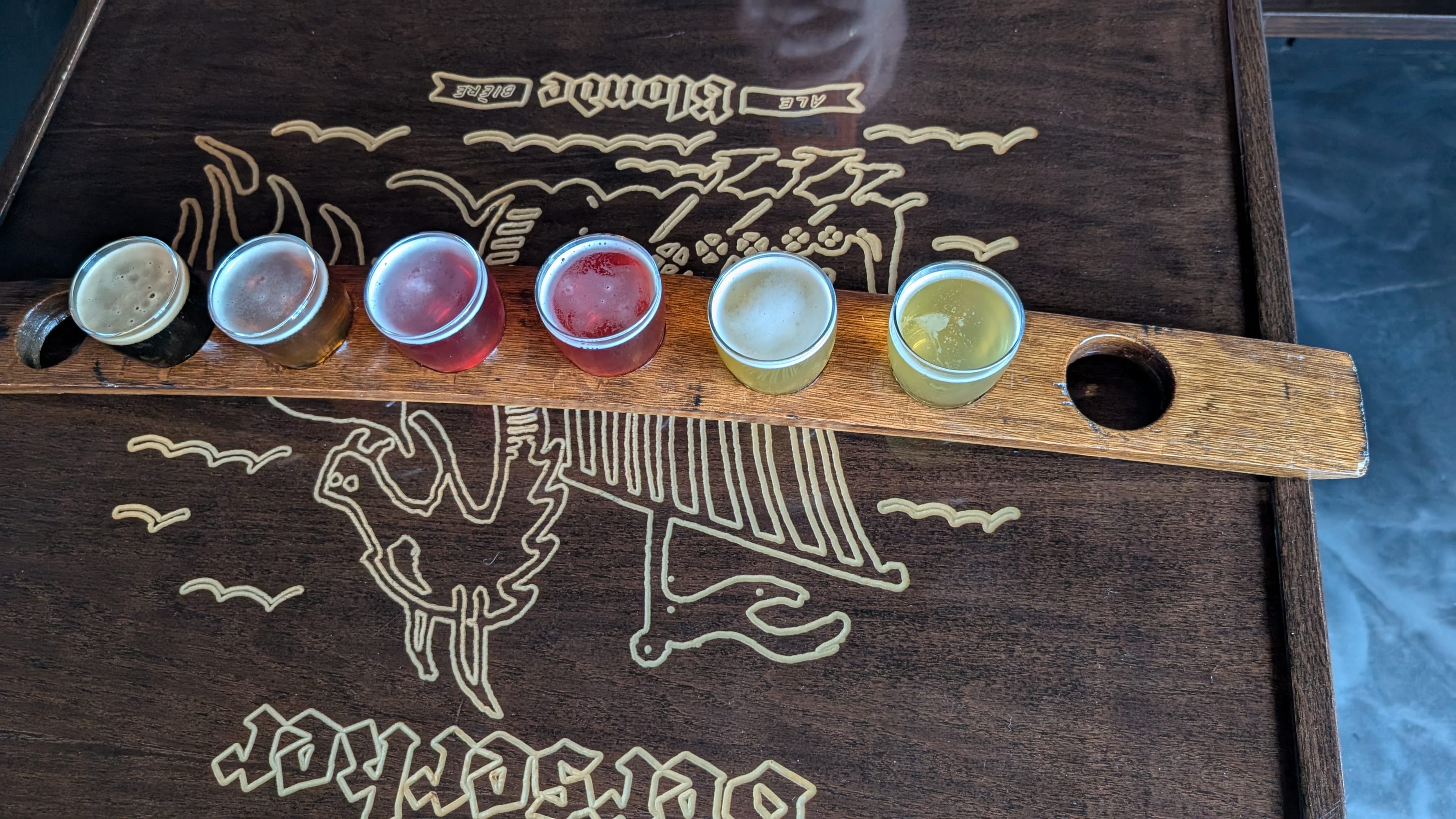 A flight of six New Level beers, from dark to pale gold, on a wooden paddle over the Berserker Blonde artwork on their table