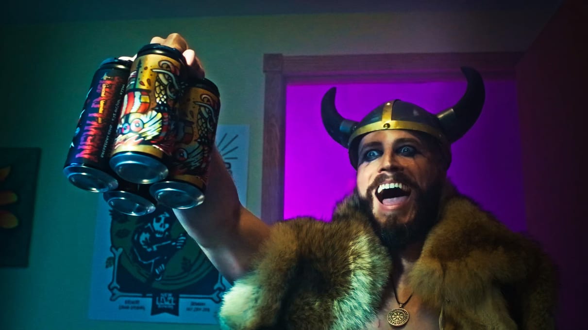 A grinning Viking in a horned helmet and fur raises a four-pack of New Level cans, from their Berserker Blonde campaign